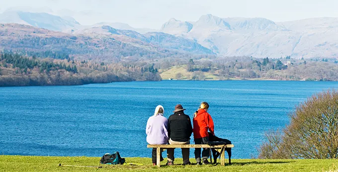 3 people sit on a bench looking over a calm blue lake on a sunny day, with steep hills in the distance. 