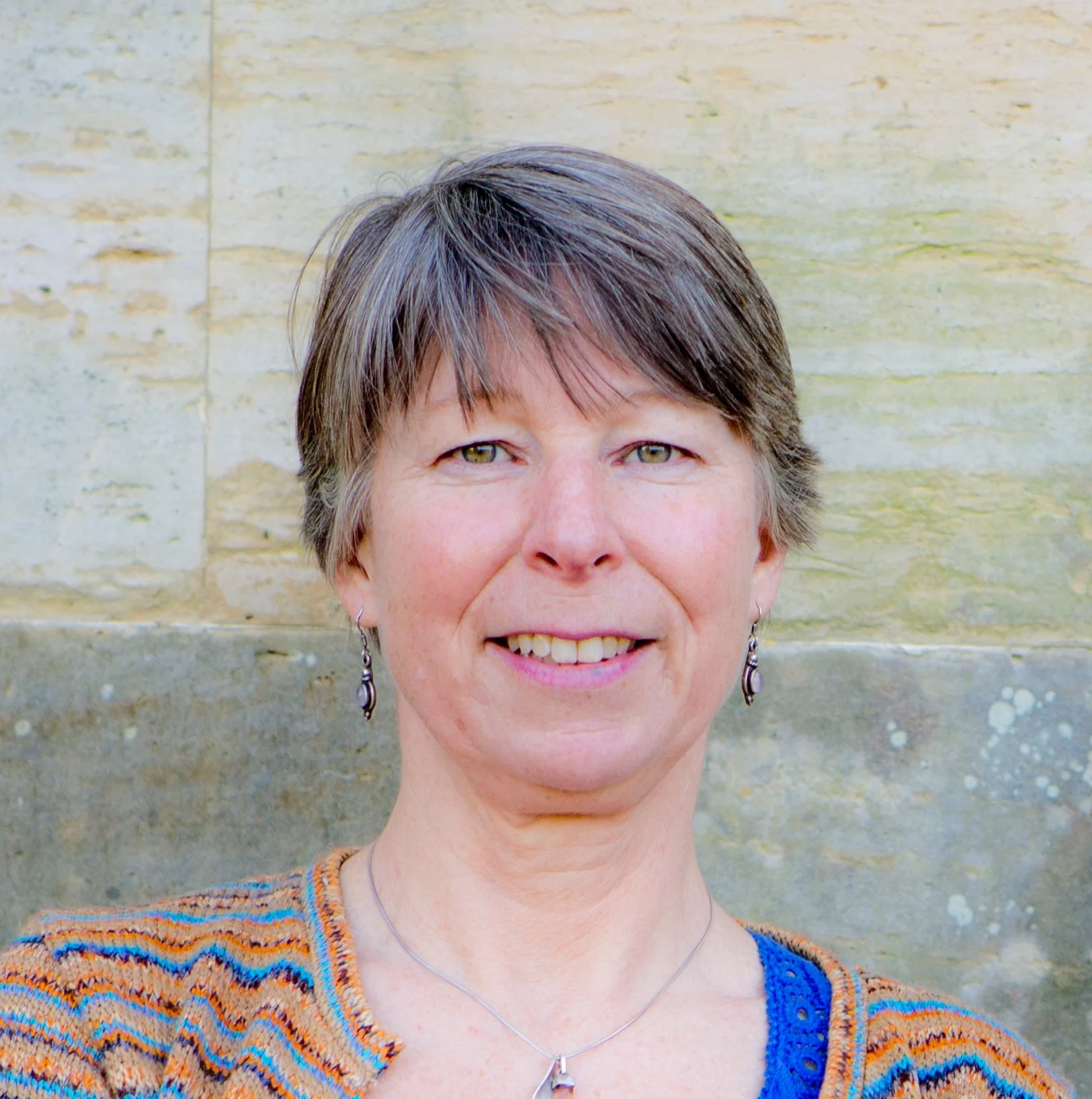 Headshot of Clare Evans, a white woman with short grey/brown hair, wearing a blue top and multicoloured striped cardigan. 