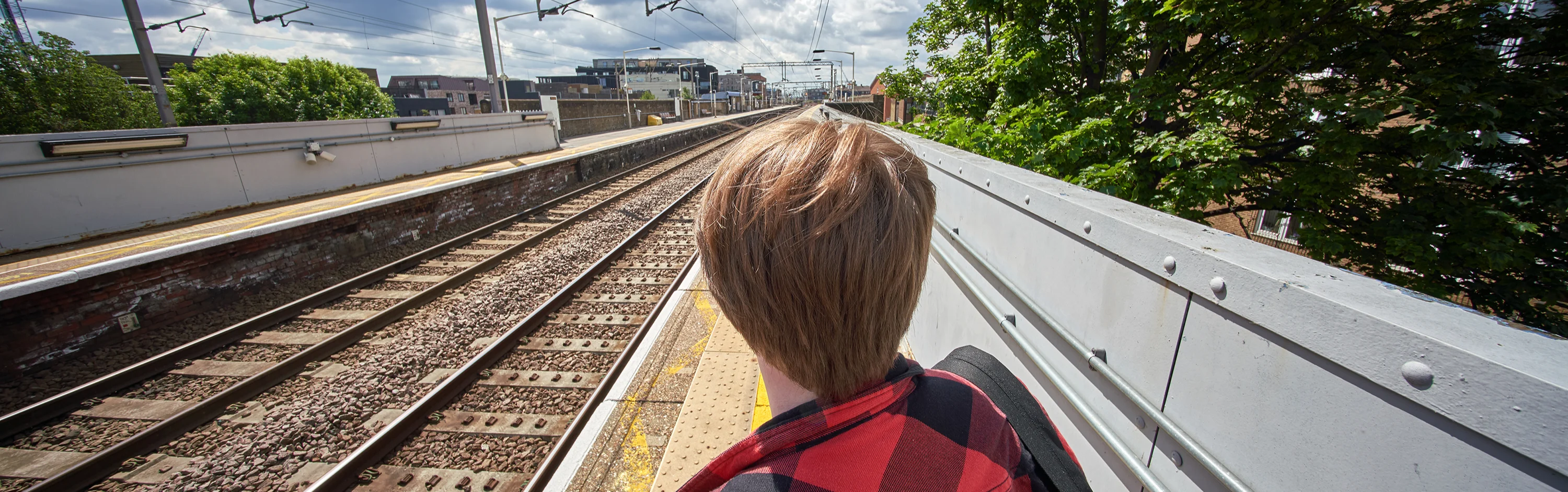 the back of a young man's head who is standing on a railway station platform in the daytime, looking into the distance down the tracks
