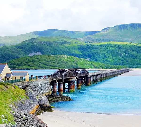 A railway bridge over a sandy beach and estuary with blue water and green hills in the background.