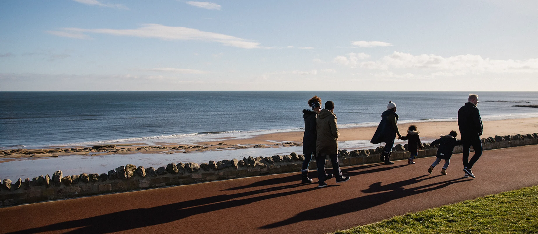 A multigenerational mixed-race family walk along a path next to a beach on a cold but sunny day