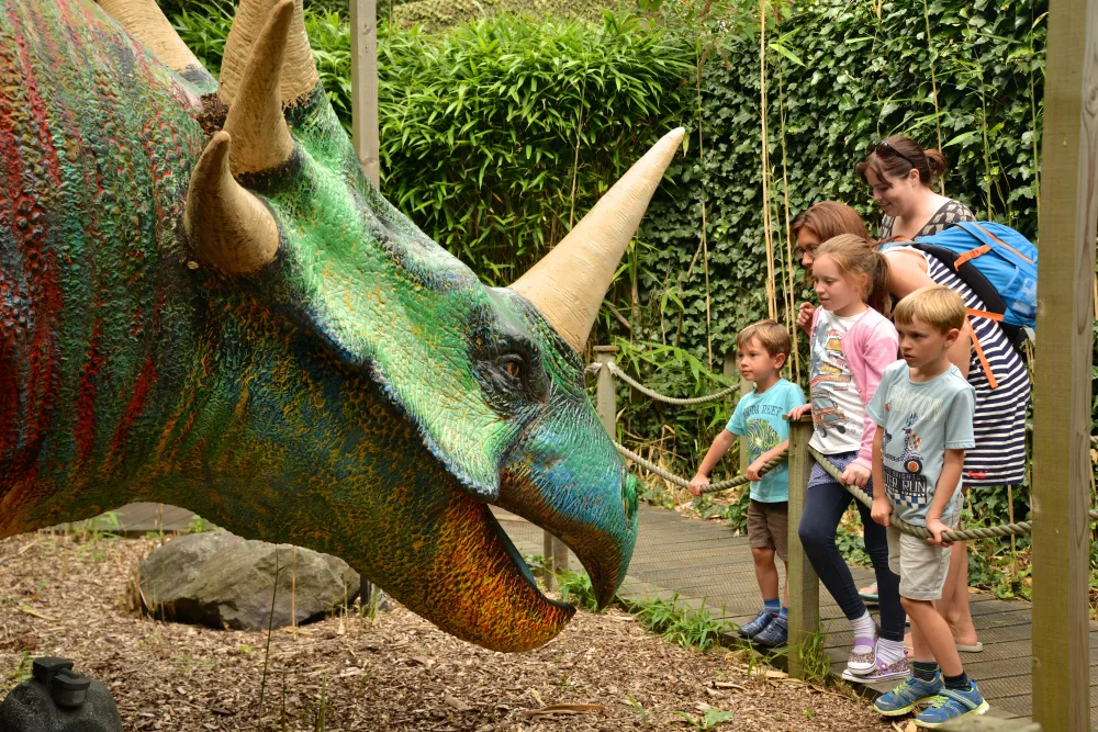 A family viewing a large, colourful triceratops dinosaur model in an outdoor exhibit surrounded by lush greenery.