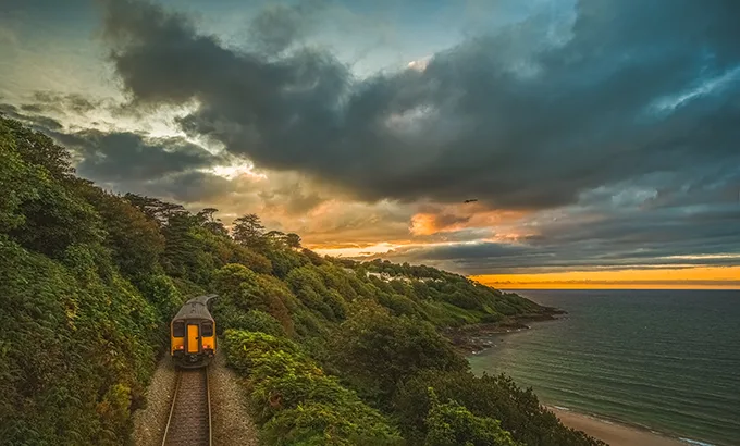 A train running along a track above a sandy beach, with trees either side of the tracks and the sun setting on the horizon. 