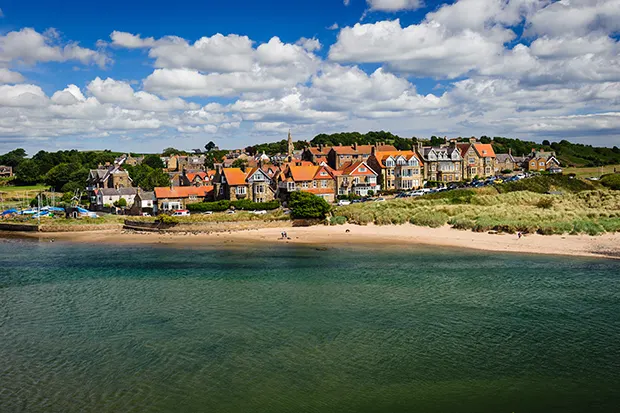 A small coastal village with red-roofed houses overlooking a sandy beach beneath a blue sky with white clouds.