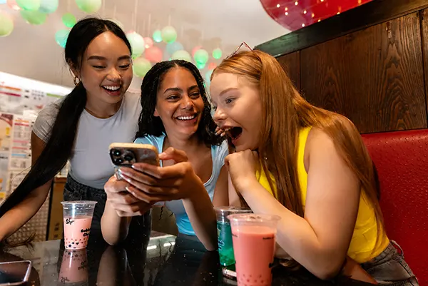 Three young women laugh and react excitedly to a phone screen in a café, with colourful bubble tea drinks on the table.