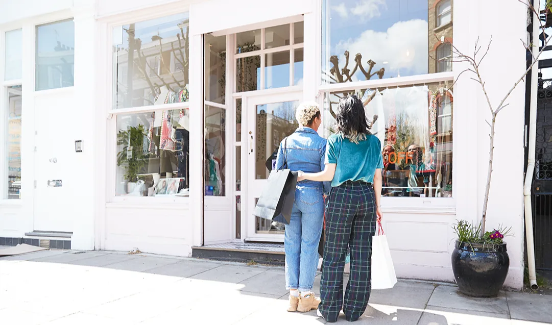 A Black woman with short bleached hair and a white woman with long dark hair, both are looking in a shop window and holding shopping bags. The white woman has her arm around the Black woman's waist. 