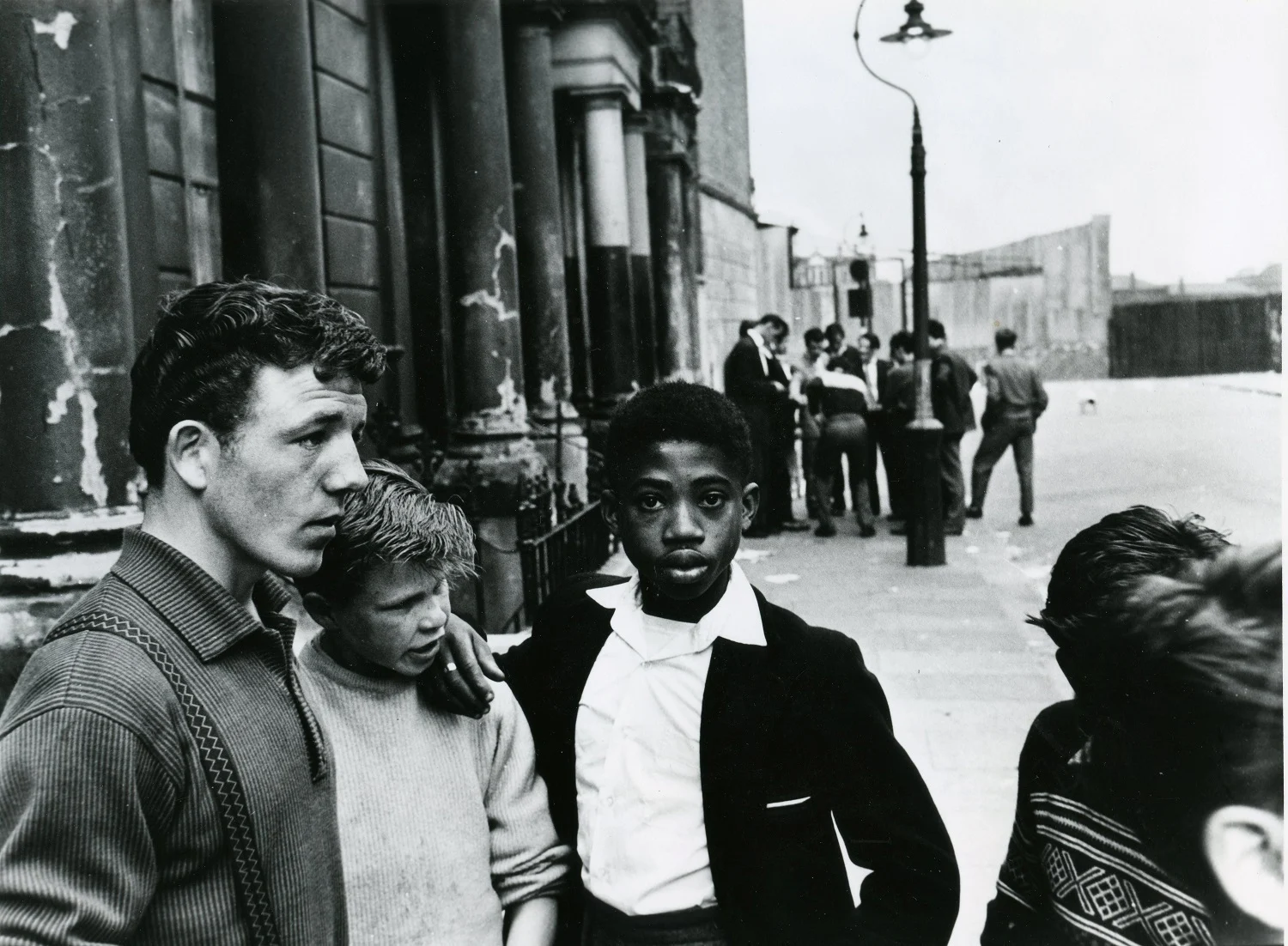 Black and white photograph of a group of Black and white young men and boys in the street in the 1950s.