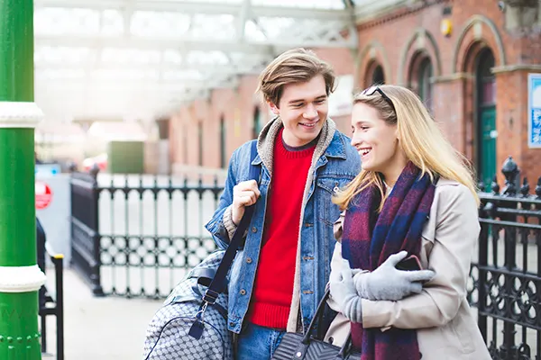 A young white man carrying a shoulder bag and a young white woman wearing gloves and a scarf, smiling and walking at a station.