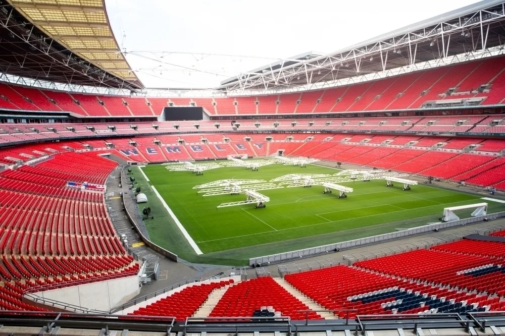 The empty interior of Wembley Stadium, with rows of red seats reaching up to the open roof around a large green pitch.