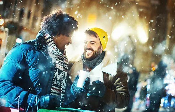 A Black man with short dreadlocks and a white man with brown hair and beard dressed up warmly and laughing together in the snow in a city street. 