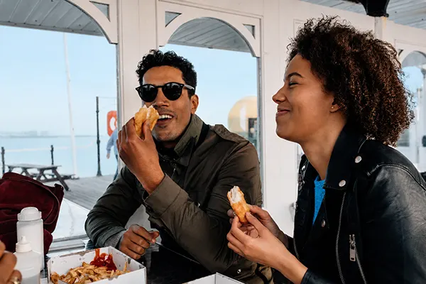 A man and woman enjoy fish and chips at a seaside café, smiling and eating together with a blue sea view behind them.