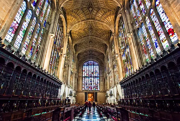 Inside a historic chapel, with high arched ceiling, large stained glass windows and dark wood seats along the side.
