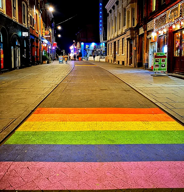 A city street at night, with large rainbow stripes painted on the ground. 