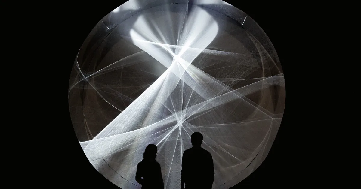 Two silhouetted figures stand before a large circular light installation with sweeping white beams against a dark background.