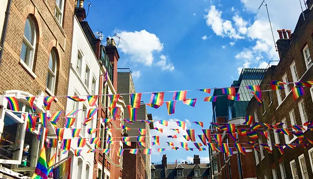 Rainbow flag bunting hanging between red brick buildings on opposite sides of street, with a blue sky in the background.