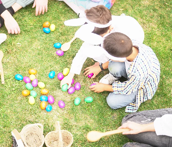 A group of children and adults seen from above, sitting on grass surrounded by colourfully wrapped eggs and wicker baskets.
