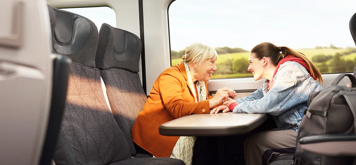 An older white woman with white hair and younger woman with long dark hair in a ponytail, sitting opposite each other at a train table and holding hands while deep in conversation, with green fields visible through the train window.  