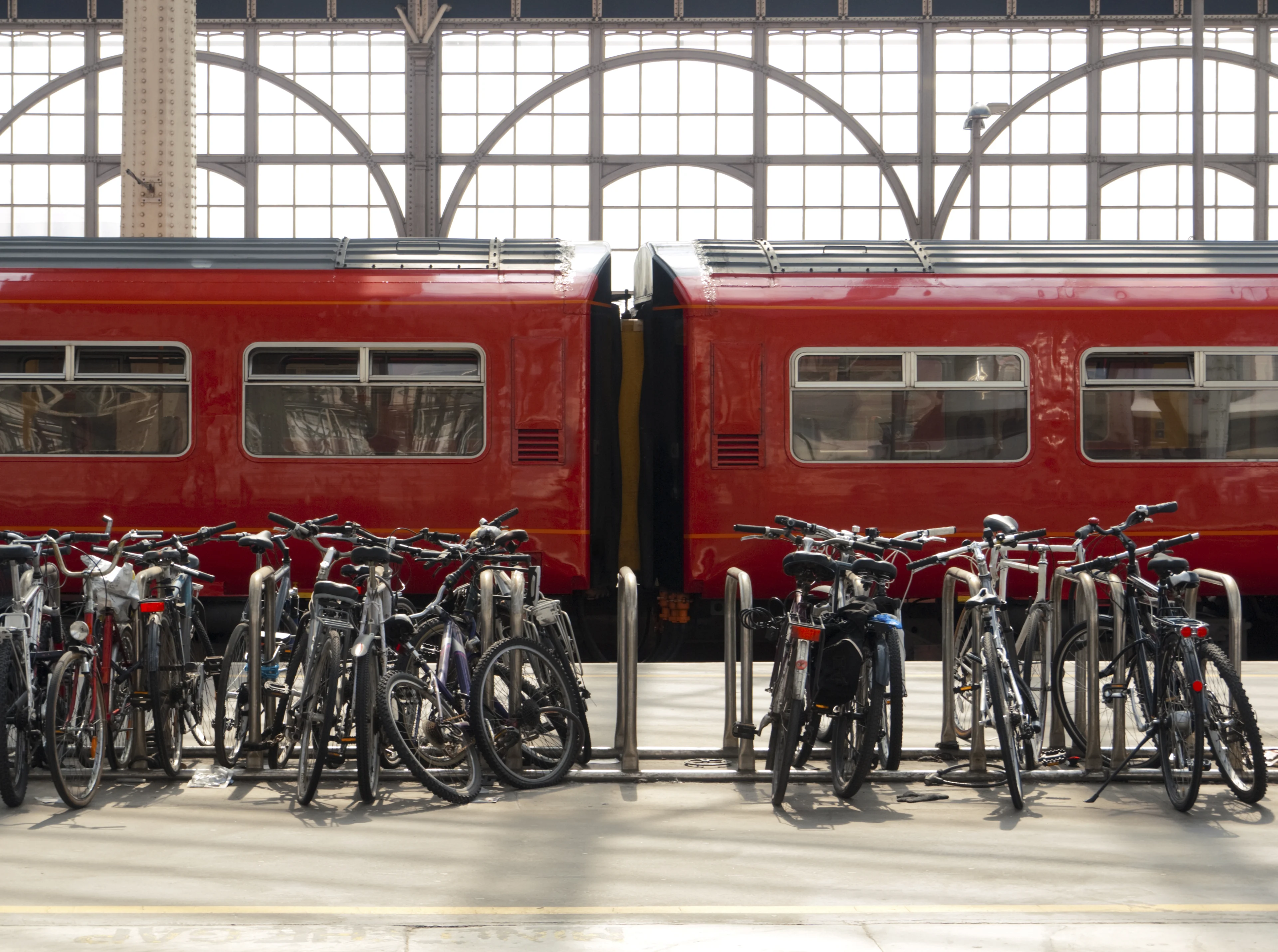 Bicycles parked at a station with a train behind them at a platform and arched windows in the background