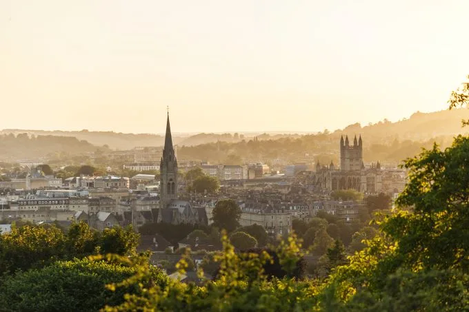 The turrets and roofs of Bath's skyline sit beneath a dusky sky with sun setting to the east. Green foliage to the foreground of the image.