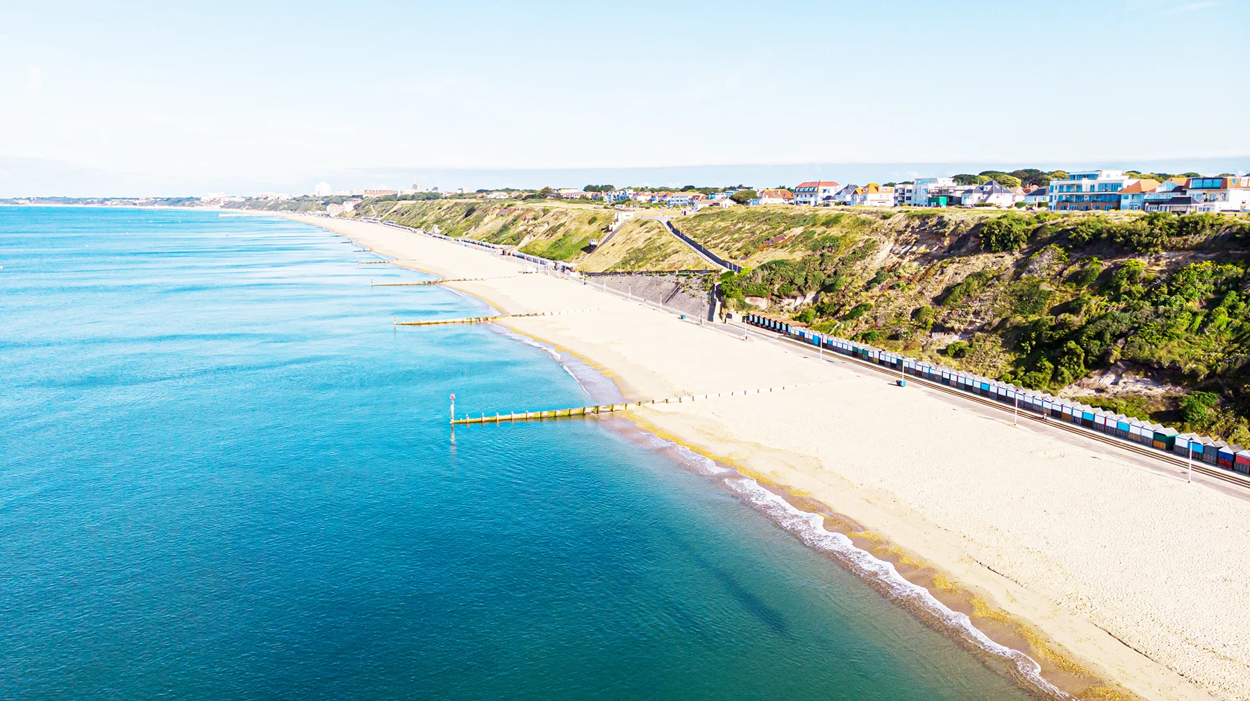 A bright blue sea and sandy beach next to a railway track with a train on it, and a row of beach huts. There is a grassy bank and houses on the other side. 