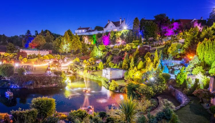 A vibrant illuminated model village and garden at night, featuring colourful lights, a pond with fountains, lush trees, and a white house on a hill.