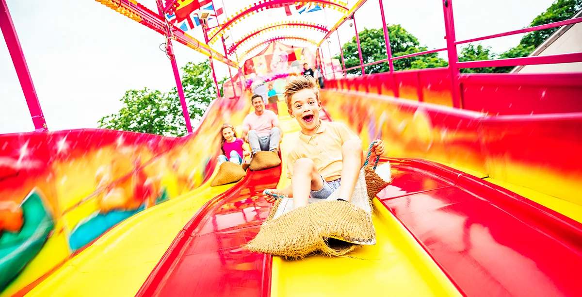 2 happy and smiling white children, and a white man close behind, sliding down a vibrant red and yellow fairground slide on burlap mats.