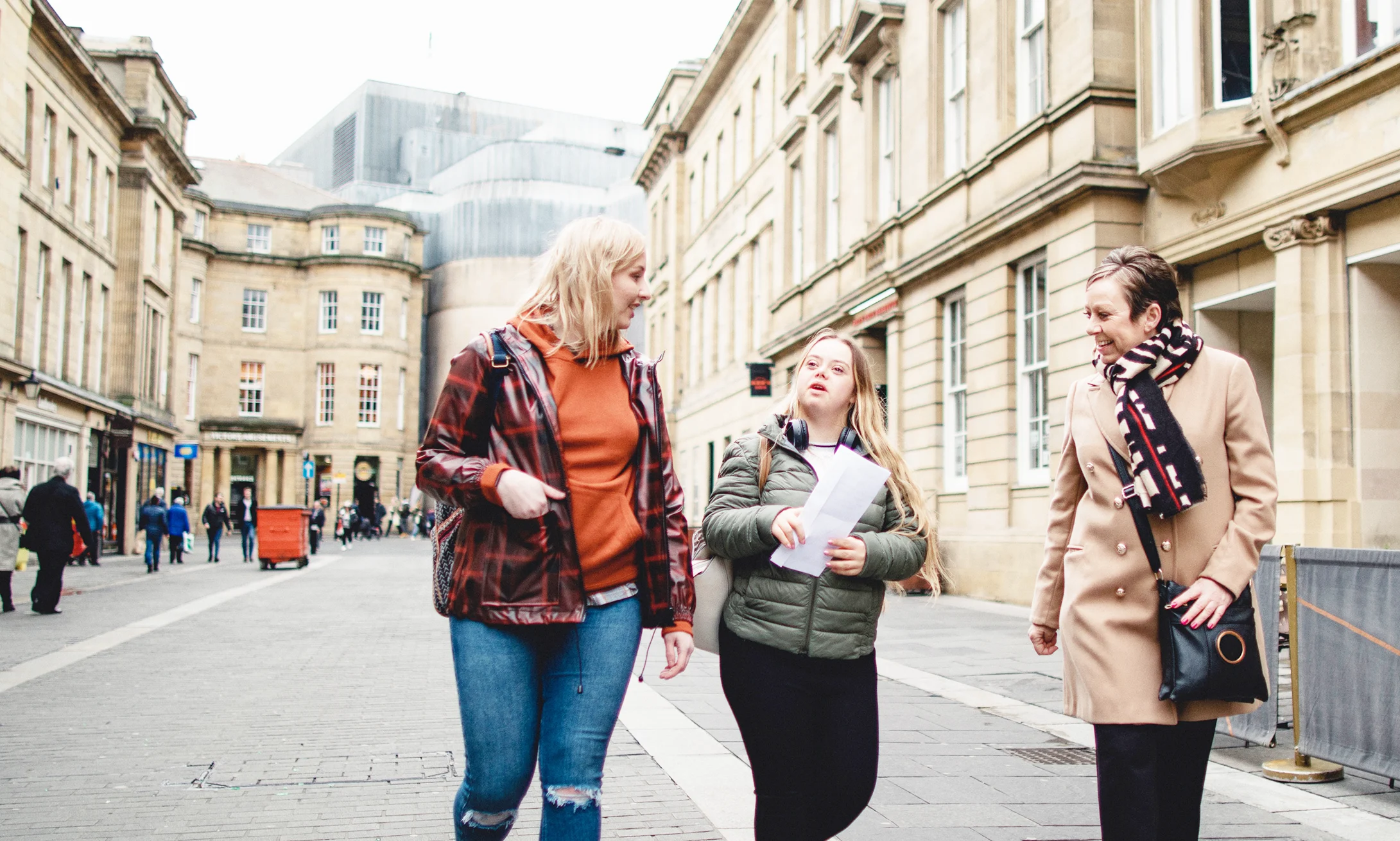 A young adult woman with Down Syndrome walking down a street in Newcastle with her friend and her mum