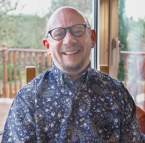 Head and shoulders image of a smiling white man with a bald heard and beard, wearing black glasses and a blue, paisley patterned shirt.