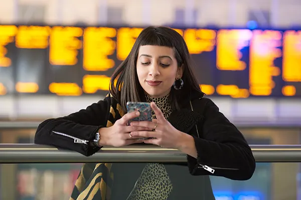 A woman with olive skin and long dark hair with a blunt fringe looks at her mobile phone screen while standing in front of a large train station departure board.