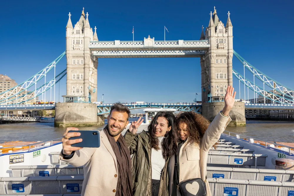 3 20-somethings pose for a selfie on a boat going under Tower Bridge on a sunny day.