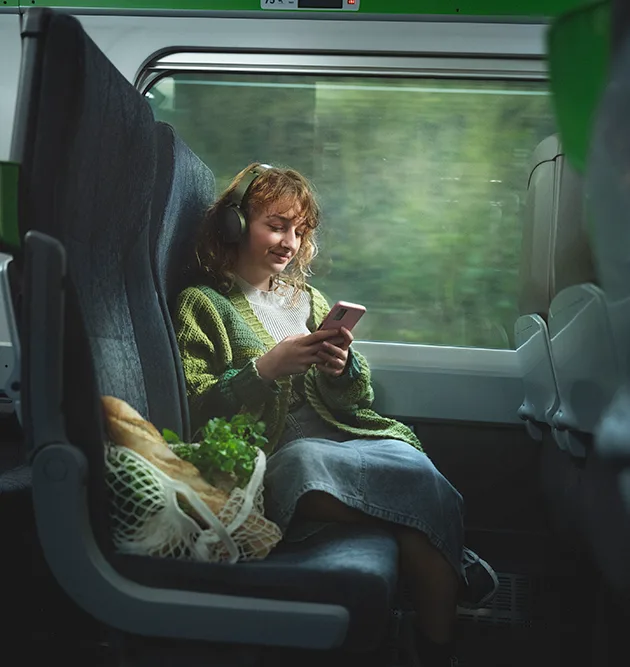 A smiling young white woman sitting on a train next to the window, she is wearing headphones and looking at her phone. 