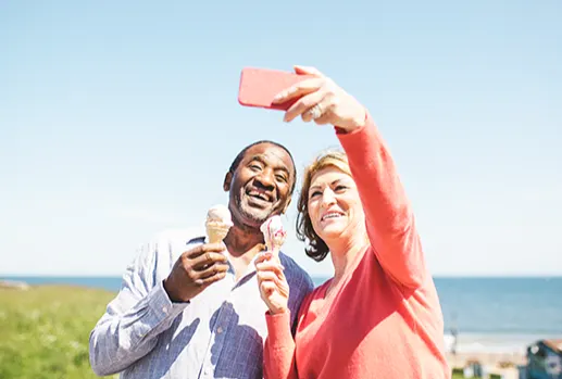 A middle aged Black man and white woman standing by a beach on a sunny day, they are both holding ice cream cones and she is taking a selfie of them.