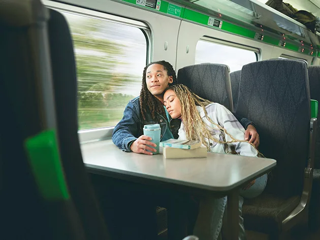 A Black man and Black woman sitting next to each other on a train, he is looking out the window and holding a travel mug while she leans on his shoulder with her eyes closed. 