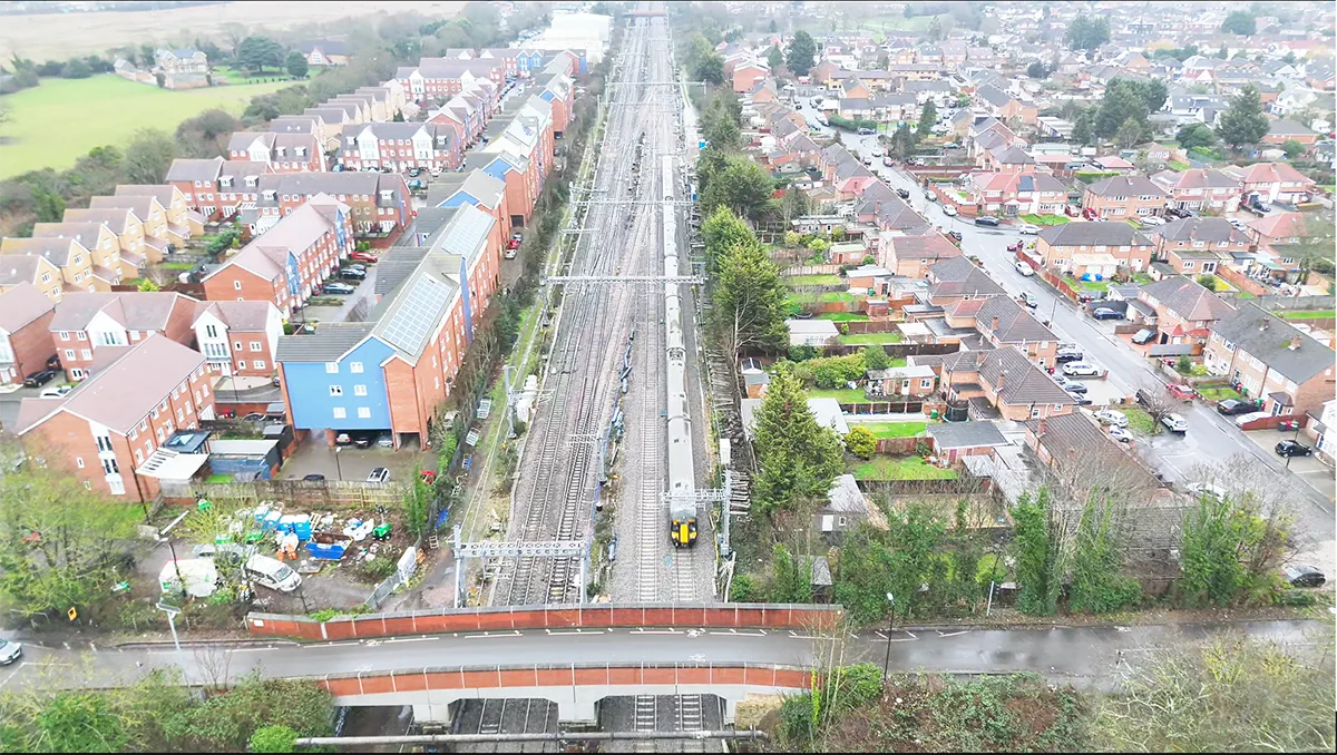 Aerial shot of a train going along tracks with buildings either side and a road bridge across. 