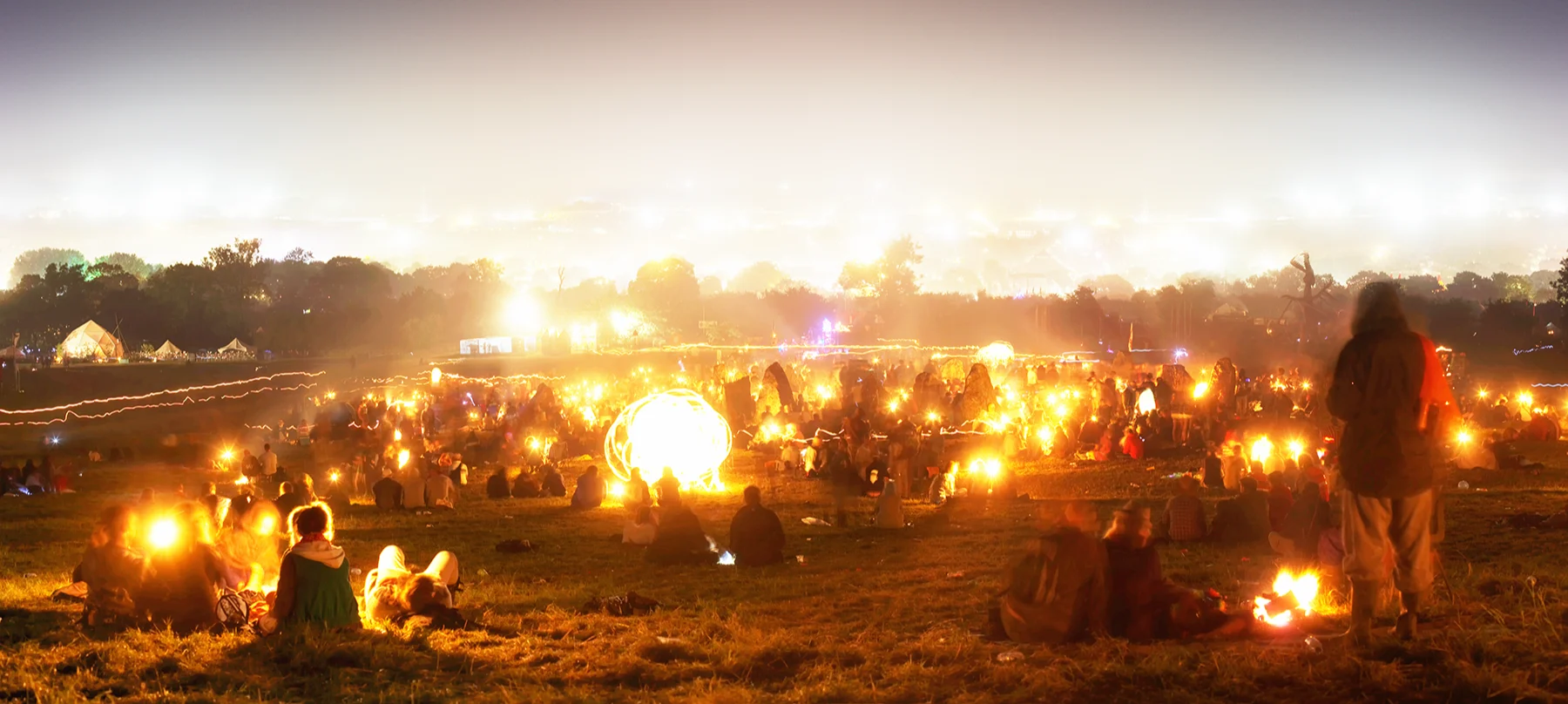 A field at dusk full of festival-goers with camp fires and the lights of Glastonbury in the distance