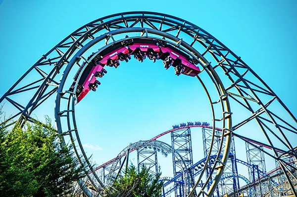 Roller coasters at a theme park with passengers suspended upside down in a bright pink carriage on a loop. 