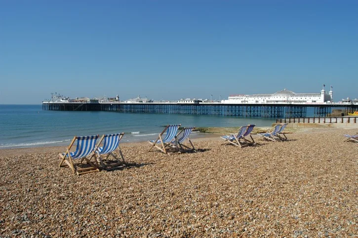 A shingle beach on a sunny day with empty deckchairs in the foreground a pier in the background.