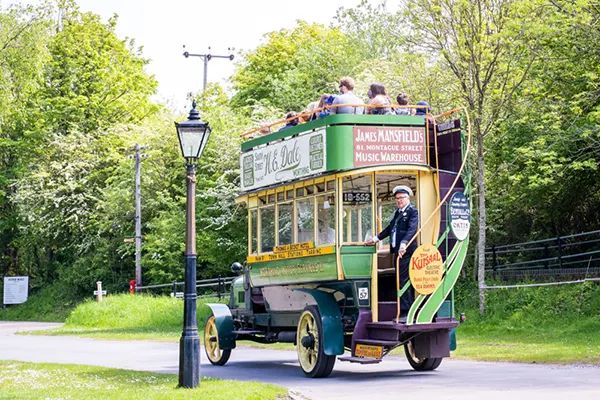 Vintage green double-decker bus travelling along a tree-lined path with passengers seated on the open top deck.
