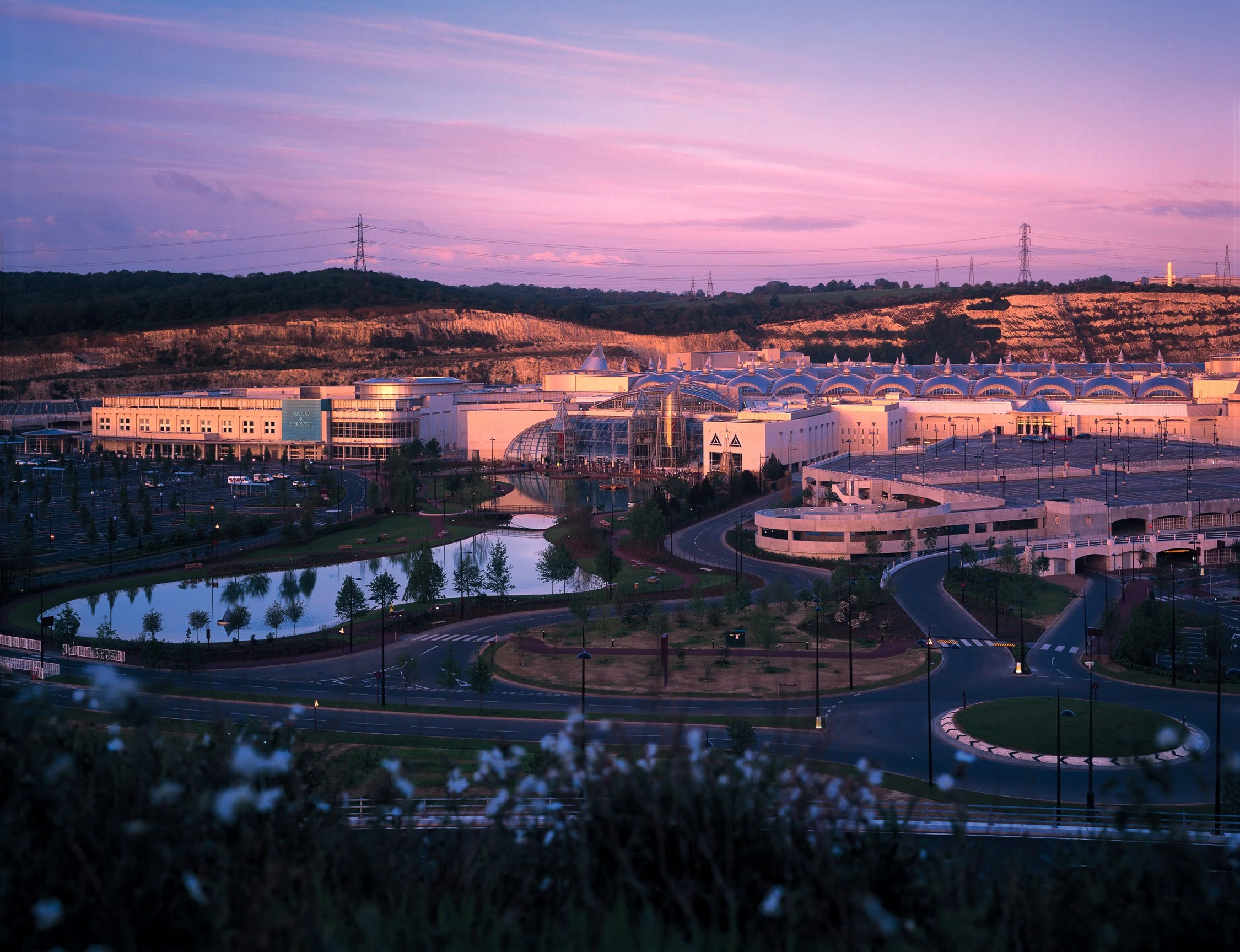 A large out-of-town shopping centre complex set by a lake and gardens, with a pink sky above. 