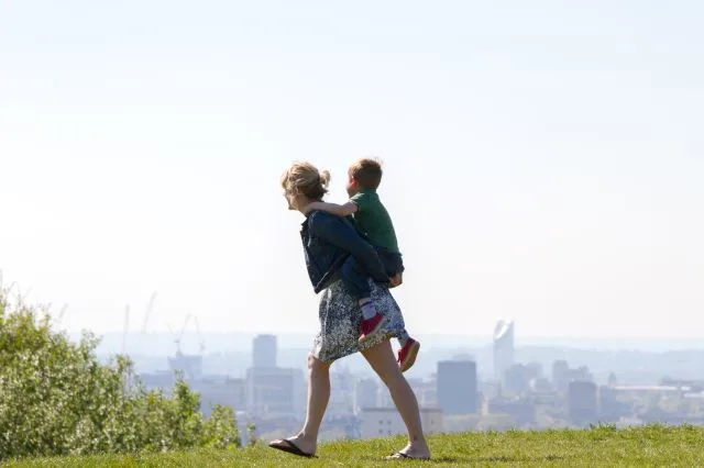 Woman walks with child on her back on grass, with city scape in the background