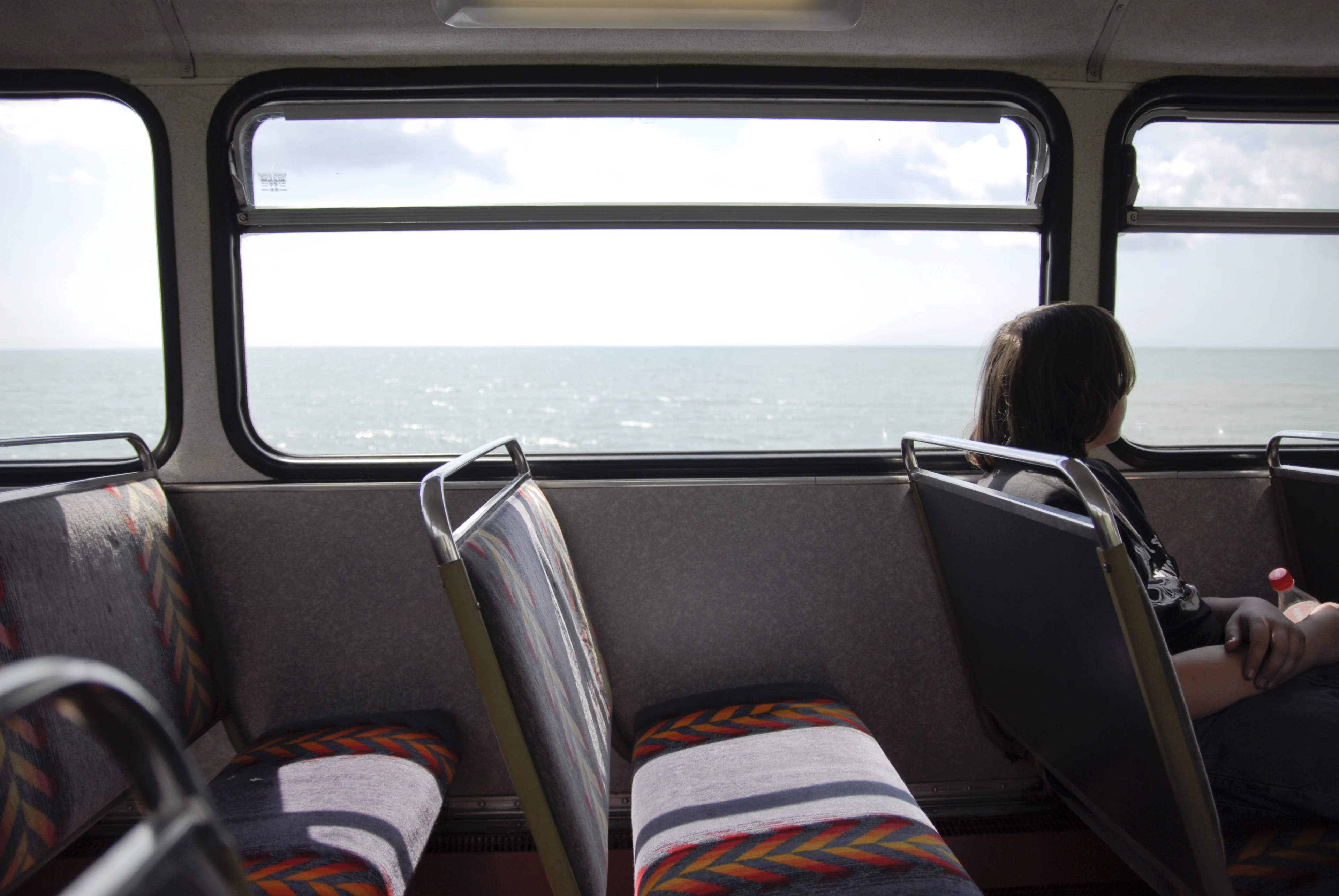 A young person alone on the top deck of a bus looks out the window out to sea on a sunny day