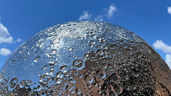 A large metallic sphere replicating the Moon photographed against a bright blue sky with scattered white clouds.