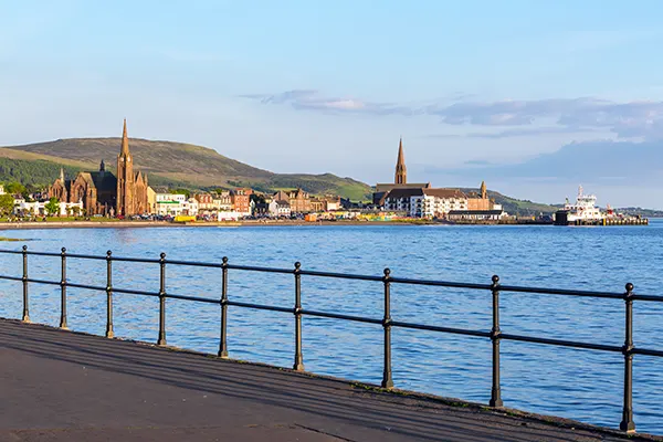 View from a seaside promenade with a metal railing looking over the water to a pretty coastal village with rolling green hills in the distance. 