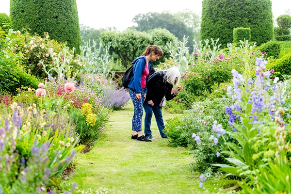 2 white women standing in a well-maintained formal garden with topiary and colourful flower beds. 1 woman is bending down to take a closer look at a flower. 