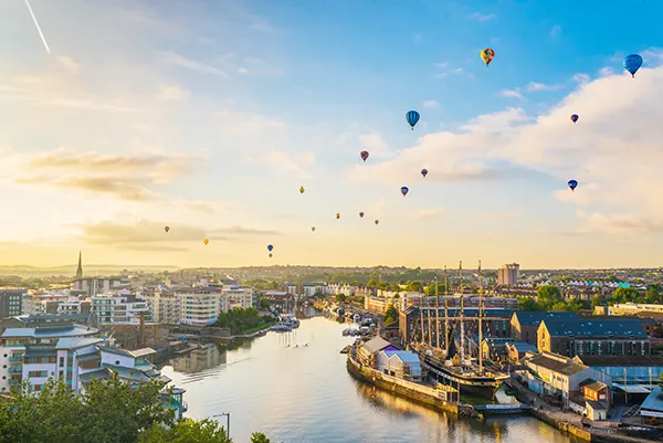 Multicoloured hot air balloons floating above a river running through a city.