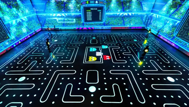 Players walking on a giant real-life reconstruction of the Pac-Man video game, with white dots and ghosts on the floor and neon lights around the walls.