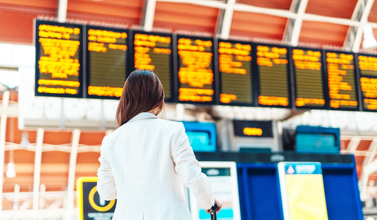 Person in white jacket looking at departure board with yellow text displays at railway station.