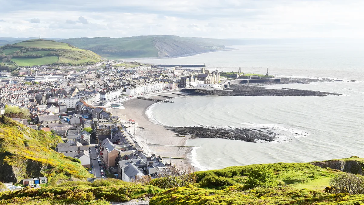 Aerial view of a coastal Welsh town with curved beach, historic buildings, and green hills overlooking the sea on a cloudy day.