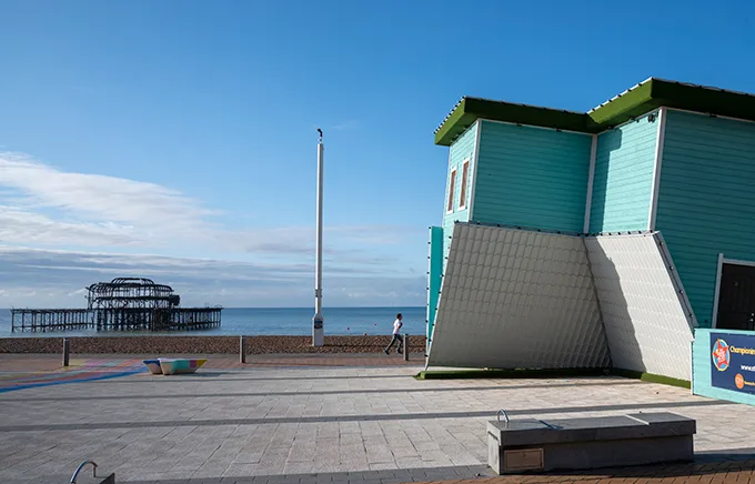 A view from the seafront at Brighton beach with an upside down house with a white roof on the ground and green wooden sides above.