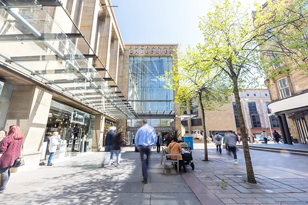 A pedestrianised city centre shopping street leading up to a large sandstone and glass shopping centre building. 